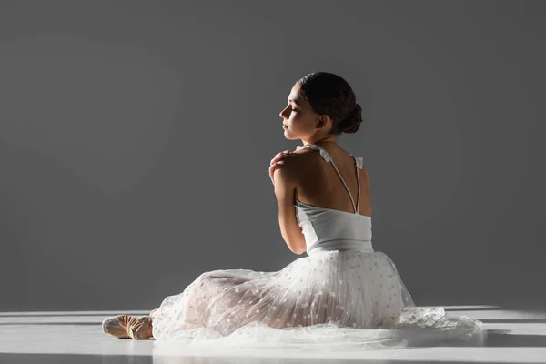 Vue latérale de la jeune ballerine touchant l'épaule sur fond gris avec lumière du soleil — Photo de stock