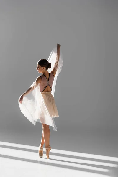 Vista lateral da bailarina elegante dançando com pano branco sobre fundo cinza com luz solar — Fotografia de Stock