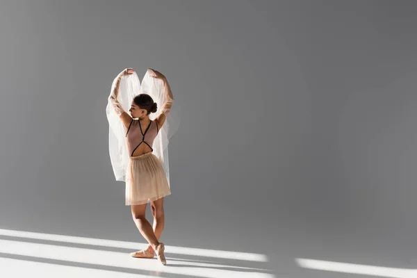 Vue latérale de la ballerine avec tissu levant les mains sur fond gris avec lumière du soleil — Photo de stock