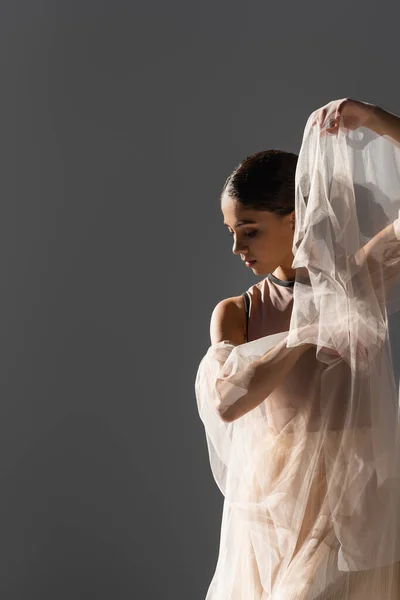 Вид сбоку танцовщицы-брюнетки, держащей в руках ткань в светлой изоляции на сером фоне — стоковое фото