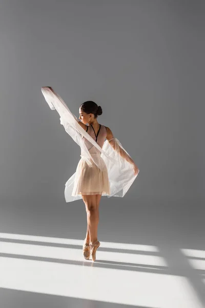 Вид збоку гарненька балерина танцює з білою тканиною на сірому фоні з сонячним світлом — стокове фото