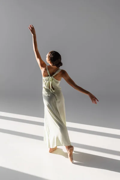 Вид на балерину в платье, поднимающую руку на сером фоне с солнечным светом — стоковое фото