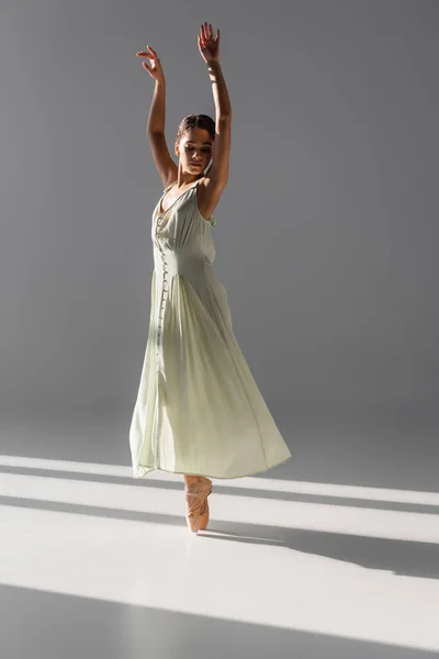 Pretty ballerina in dress spinning on grey background with sunlight — Stock Photo