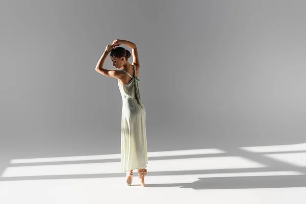 Vista lateral de la bailarina morena en zapatos puntiagudos y vestido sobre fondo gris con luz solar — Stock Photo