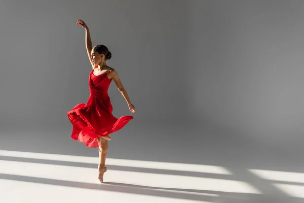 Vista lateral de la joven bailarina en vestido rojo bailando sobre fondo gris con luz solar - foto de stock