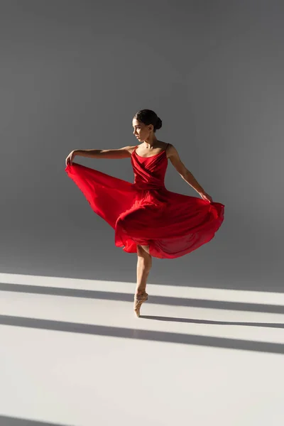 Vue latérale de la ballerine dansant et tenant une robe rouge sur fond gris avec lumière du soleil — Photo de stock