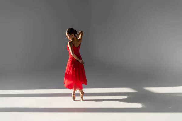 Vista lateral de la bailarina en vestido rojo y zapatos puntiagudos sobre fondo gris con luz solar - foto de stock