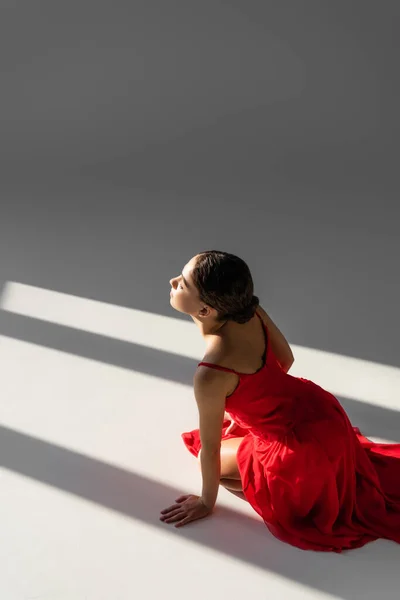 Високий кут зору танцюриста в червоній сукні, що сидить на сірому фоні з сонячним світлом — стокове фото
