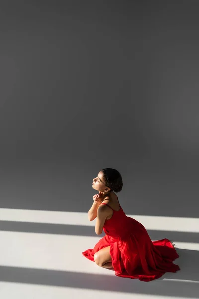 Vue latérale de la danseuse brune en robe rouge assise sur fond gris avec lumière du soleil — Photo de stock