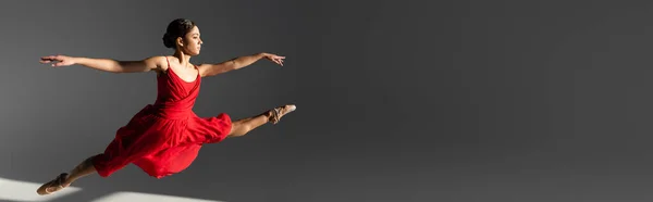 Seitenansicht der brünetten Ballerina im roten Kleid, die auf grauem Hintergrund mit Sonnenlicht springt, Banner — Stockfoto