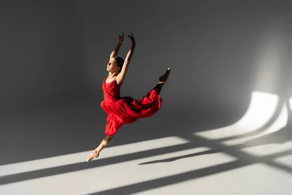 Side view of pretty ballerina in red dress jumping and raising hands on grey background with sunlight — Stock Photo