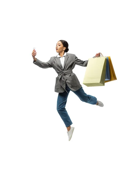 Bailarina olhando para smartphone enquanto segurando sacos de compras e pulando isolado no branco — Fotografia de Stock