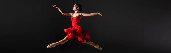 Side view of ballerina in pointe shoes and red dress jumping isolated on black, banner — Stock Photo