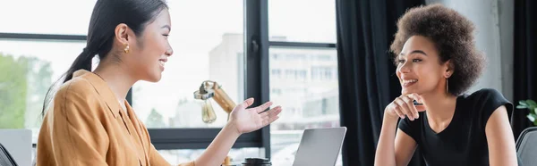 Asian businesswoman pointing with hand while talking to smiling african american colleague, banner — Stock Photo