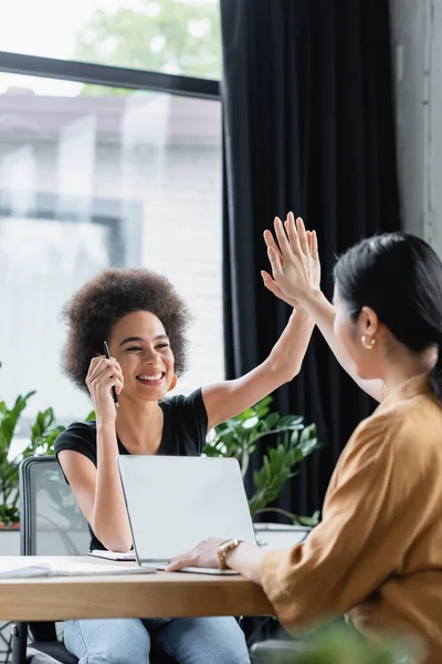 Heureux interracial femmes d'affaires donnant haute cinq tout en travaillant près d'ordinateur portable sur le lieu de travail — Photo de stock