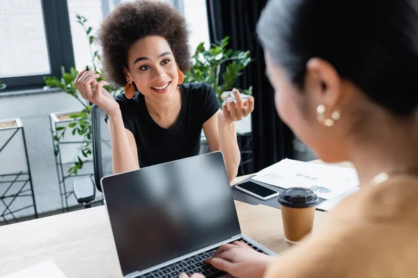 Cheerful african american woman talking to blurred colleague near laptop with blank screen — Stock Photo