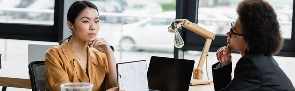 Thoughtful asian designer showing clipboard with sketch to african american colleague, banner — Stock Photo