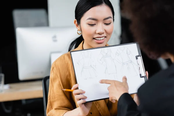 Borrosa mujer de negocios apuntando a dibujo en manos de sonriente asiático diseñador - foto de stock