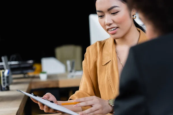 Asiatische Geschäftsfrau zeigt mit Bleistift auf Klemmbrett neben verschwommenem afrikanisch-amerikanischem Kollegen — Stockfoto