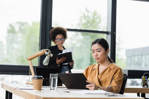 Asian businesswoman looking at clipboard near african american manager standing by window — Stock Photo