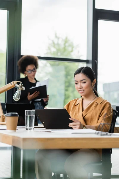 Lächelnde asiatische Geschäftsfrau beim Blick auf Klemmbrett neben verschwommenem afrikanisch-amerikanischem Kollegen am Fenster — Stockfoto