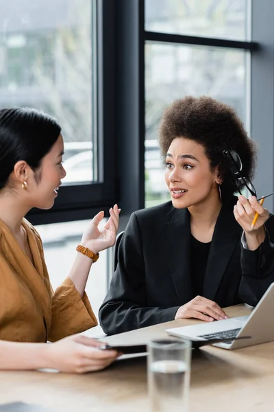 Asiático gerente gesto mientras habla sonriendo y sorprendido africano americano mujer de negocios con gafas - foto de stock