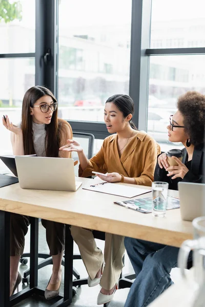 Asiatische Geschäftsfrau zeigt auf Laptop, während sie mit multiethnischen Kollegen im Büro spricht — Stockfoto
