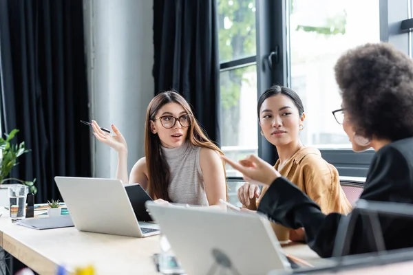 Empresarias multiculturales haciendo gestos mientras hablan cerca de computadoras en la oficina - foto de stock