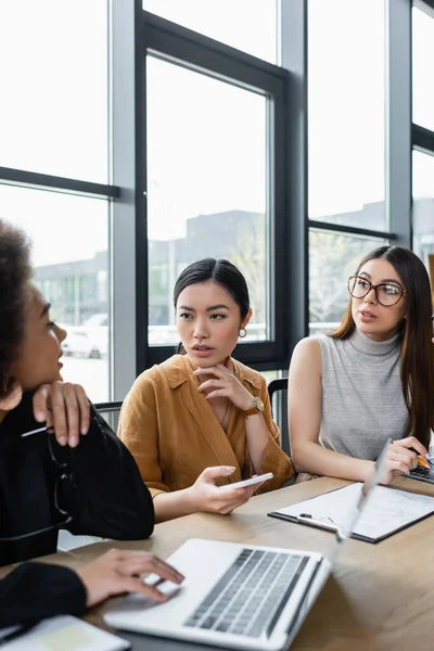 Borrosa mujer de negocios afroamericana hablando con colegas interracial cerca de la computadora portátil en la oficina — Stock Photo