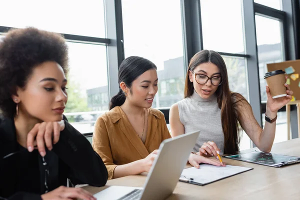 Businesswoman pointing with pencil at clipboard near smiling asian colleague and blurred african american manager — Stock Photo