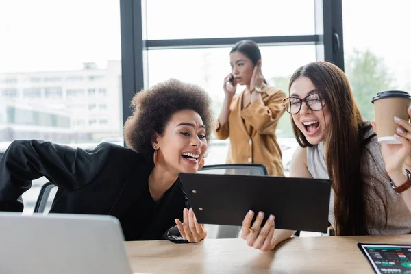 Excitado interracial businesswomen mirando portapapeles cerca asiático colega hablando en smartphone en borrosa fondo - foto de stock