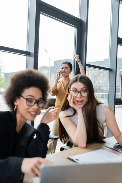 Emocionado asiático mujer de negocios hablando en smartphone cerca sorprendido interracial colegas en borrosa primer plano - foto de stock