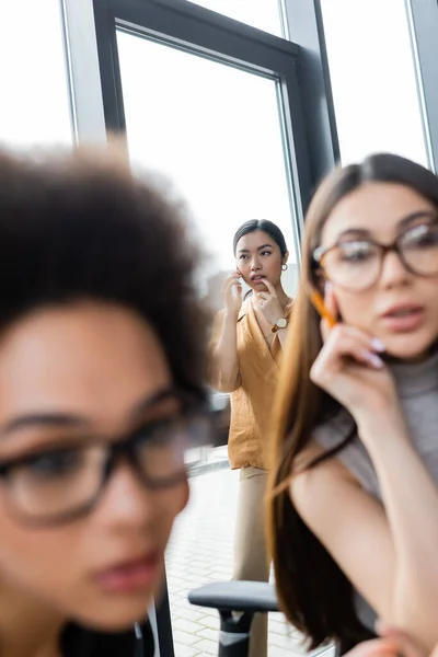 Asiático mujer de negocios hablando en smartphone cerca ventana y borrosa interracial managers - foto de stock