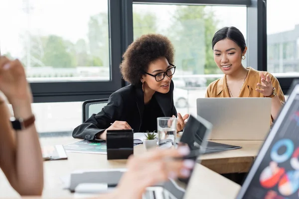 Positivo asiático mujer de negocios apuntando a portátil mientras se trabaja con africano americano colega - foto de stock