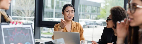 Smiling asian woman pointing with hand while talking to blurred multiethnic colleagues, banner — Stock Photo