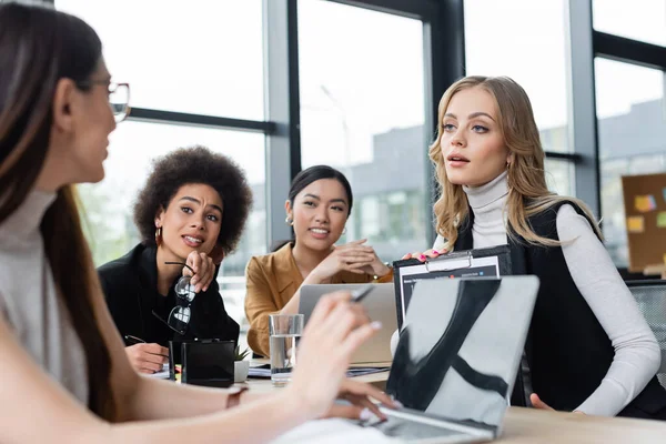 Mujer de negocios borrosa apuntando a la computadora portátil mientras trabajaba con colegas multiétnicos - foto de stock