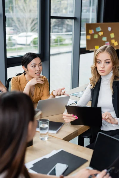 Asian businesswoman talking to colleagues near laptops and clipboards at workplace — Stock Photo