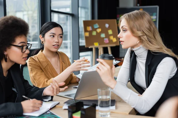 Asiático mujer de negocios apuntando con mano cerca de portátil durante la discusión con interracial colegas — Stock Photo