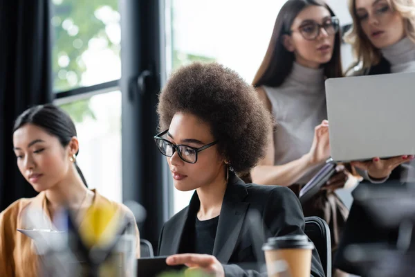 Joven mujer de negocios afroamericana en gafas que trabajan en la oficina cerca de colegas borrosos - foto de stock