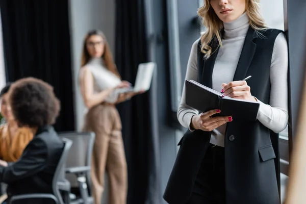 Young businesswoman writing in notebook near blurred multicultural coworkers — Stock Photo