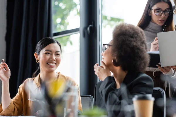 Joyeuse asiatique femme d'affaires souriant près de collègues multiethniques travaillant dans le bureau — Photo de stock