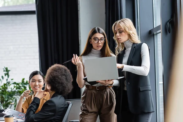 Donne d'affari bionde e brune che parlano vicino al laptop e ai colleghi multiculturali in ufficio — Foto stock