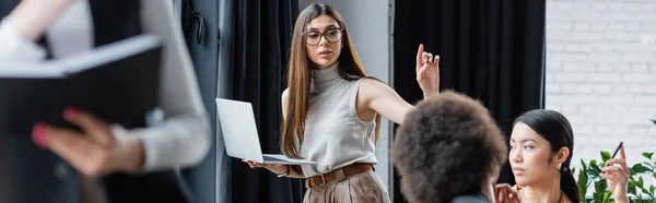 Donna d'affari con computer portatile che punta con dito vicino a manager multietnici offuscati, banner — Foto stock