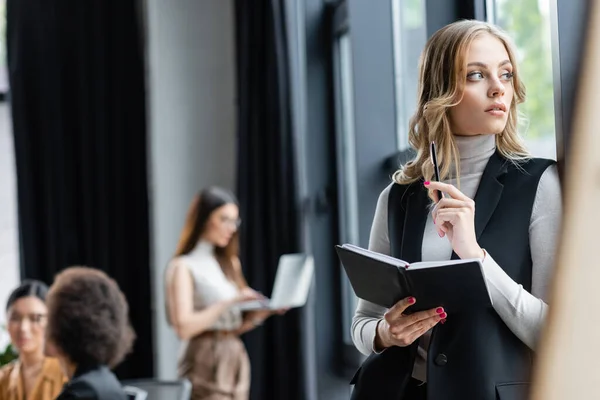 Pensativa mujer de negocios con cuaderno y pluma mirando hacia otro lado cerca borrosa multicultural compañeros de trabajo - foto de stock