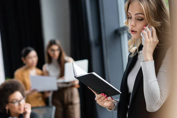 Young businesswoman looking in notebook while talking on cellphone near blurred multiethnic colleagues — Stock Photo