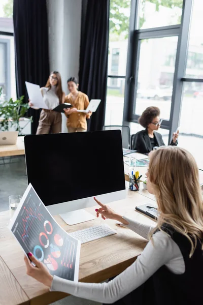 Blonde businesswoman with infographics pointing at computer monitor with blank screen in office — Stock Photo