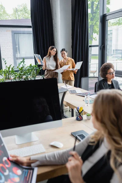 Verschwommene Geschäftsfrau blickt auf Monitor mit leerem Bildschirm, während interrassische Kollegen am Hintergrund arbeiten — Stockfoto