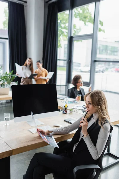 Pensativa mujer de negocios mirando infografías cerca del monitor de la computadora y colegas borrosos - foto de stock