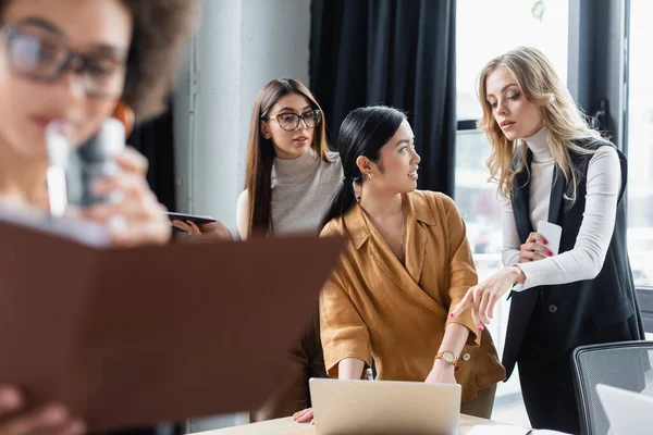 Rubia mujer de negocios con smartphone apuntando a portátil cerca asiático colega - foto de stock