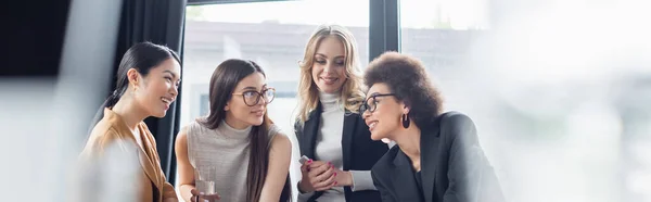 Cheerful multicultural businesswomen talking in office on blurred foreground, banner — Stock Photo